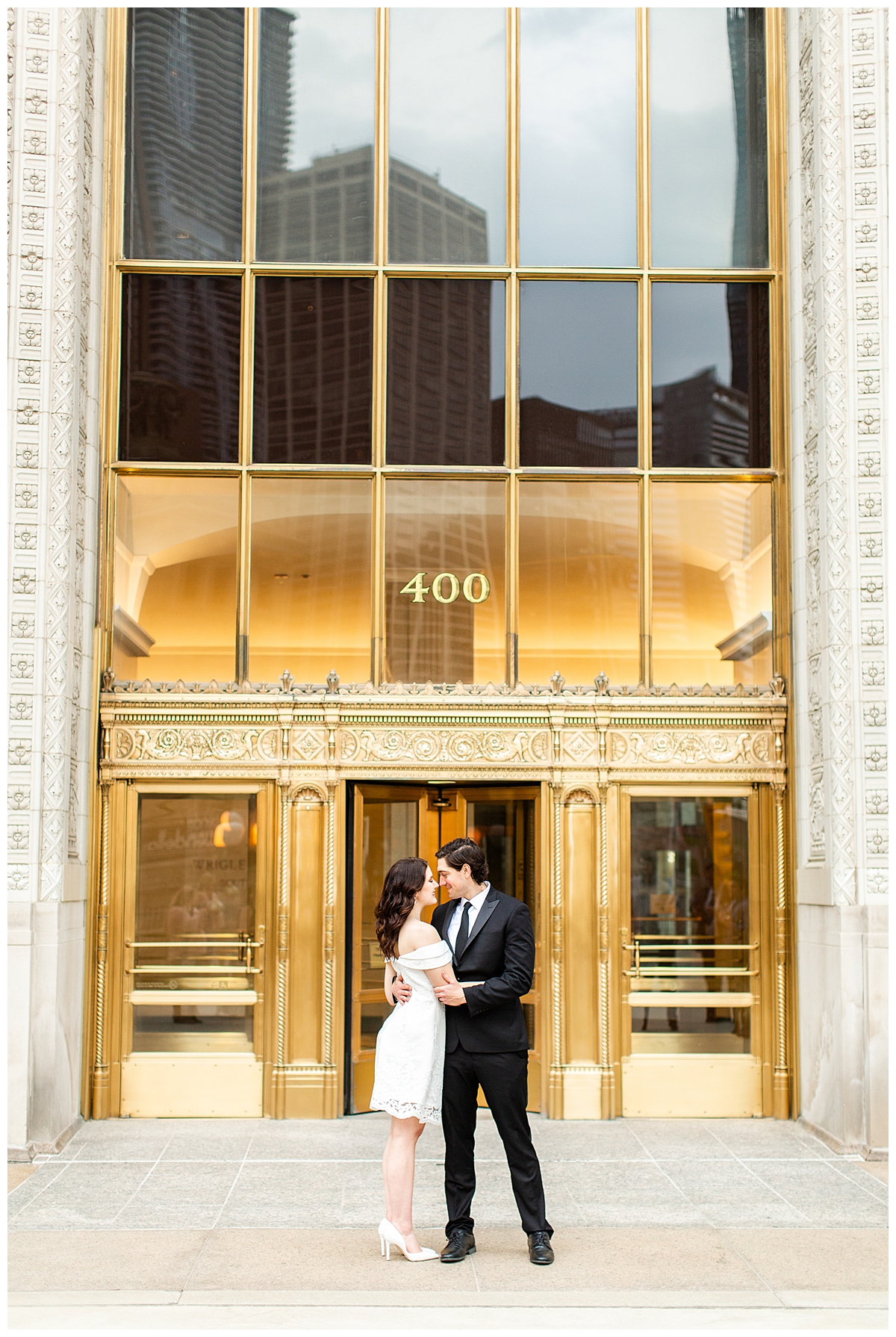 A couple takes an engagement portrait outside of the golden doors at the Wrigley Building in Chicago. They are dressed very formally. 