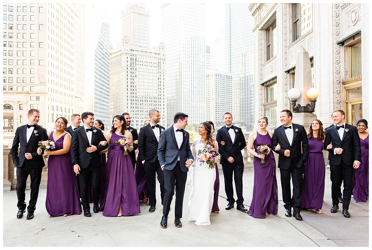 A wedding party takes a portrait outside of the Wrigley Building in Chicago. The couple is dressed whimsically and the bridesmaids are wearing purple. 