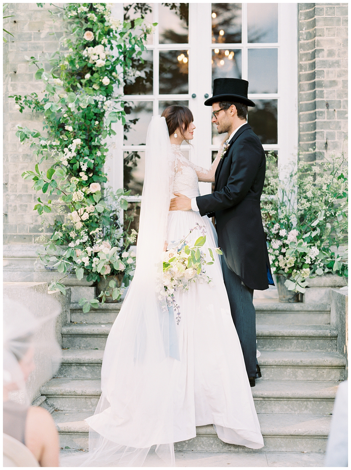 A couple poses for a wedding portrait on a set of steps in front of a floral altar. They are dressed in black tie and he is wearing a top hat. 