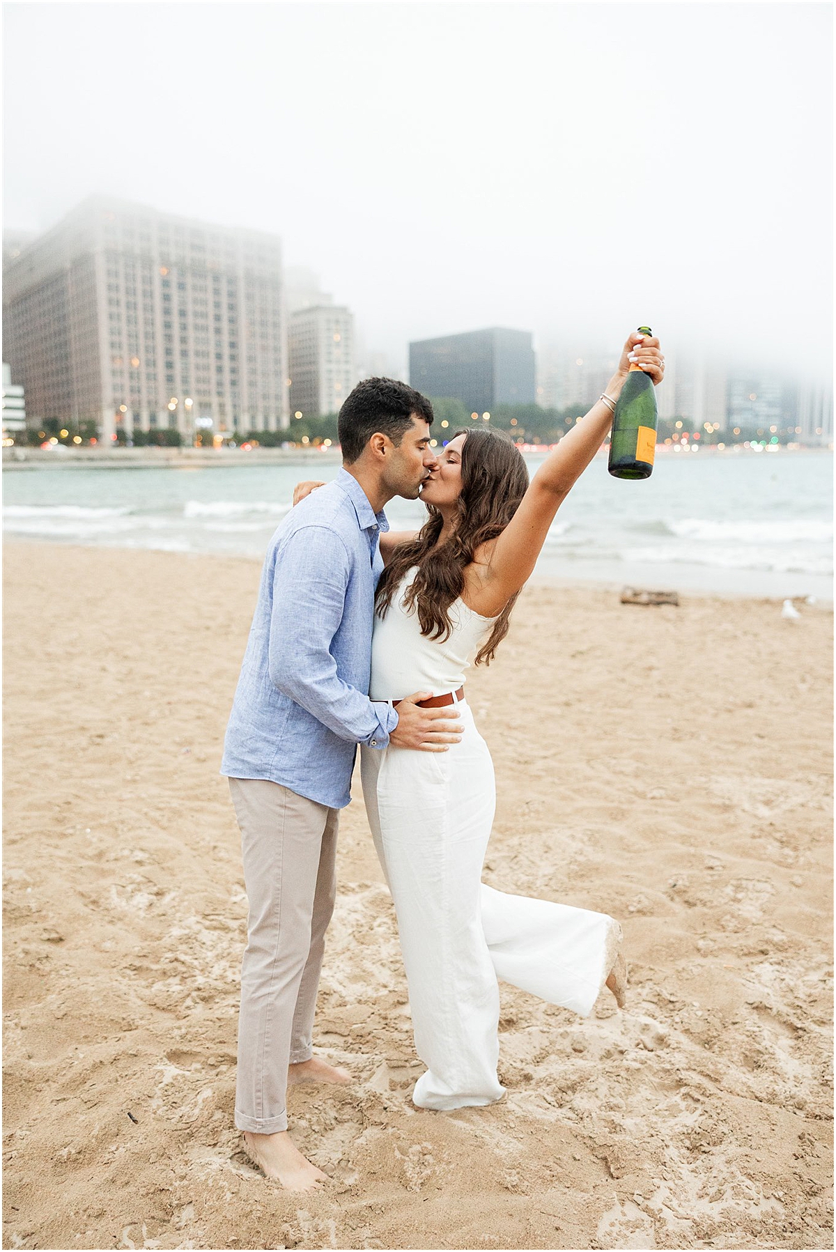 engaged couple in neutral outfits kiss at North Avenue Beach while woman holds bottle of champagne