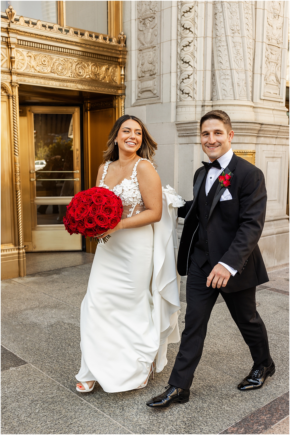 bride holding red rose wedding bouquet as she walks with groom outside of Wrigley Building in Chicago