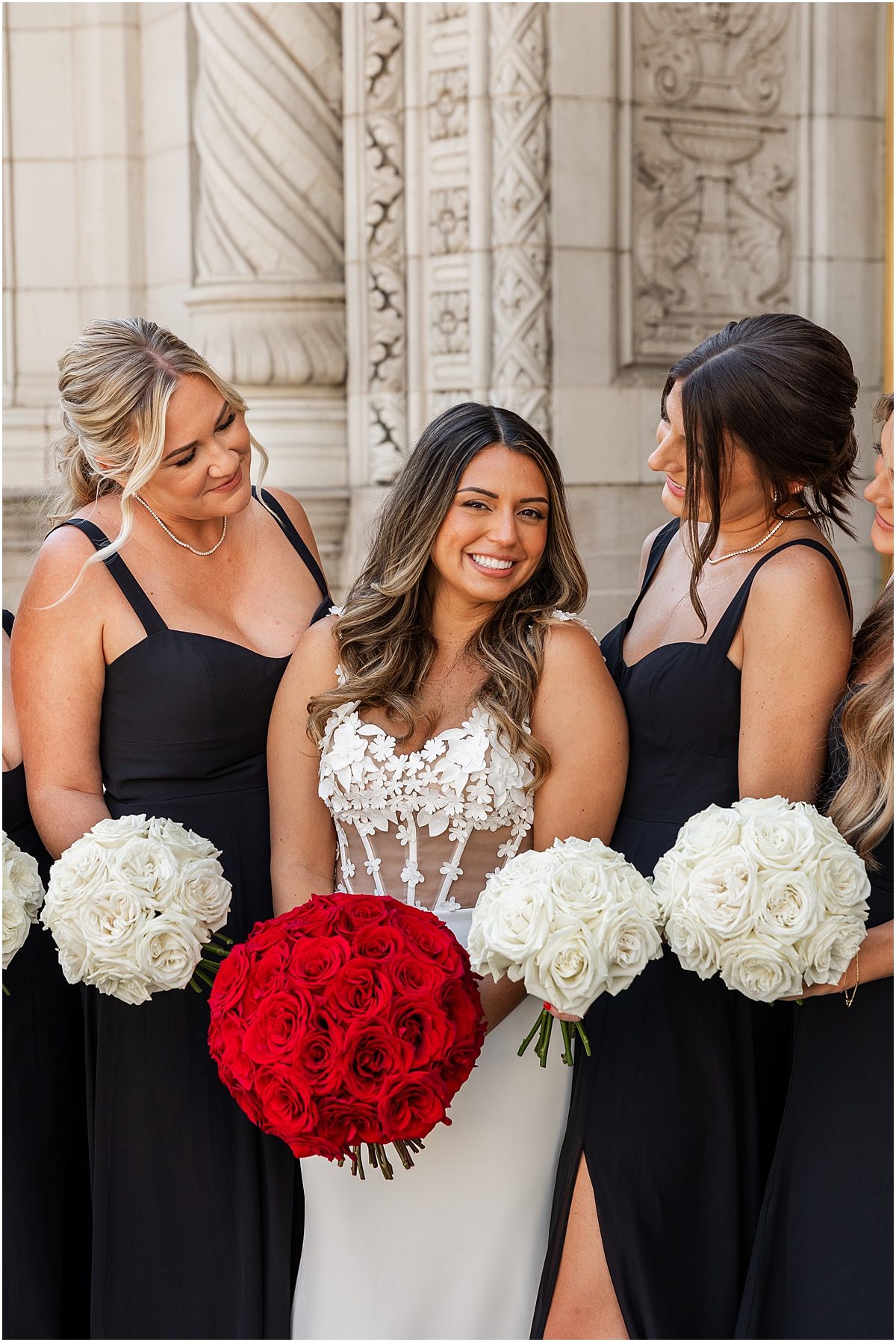 bride in corset style wedding dress holding red rose bouquet with bridesmaids in black dresses and holding white rose bouquets