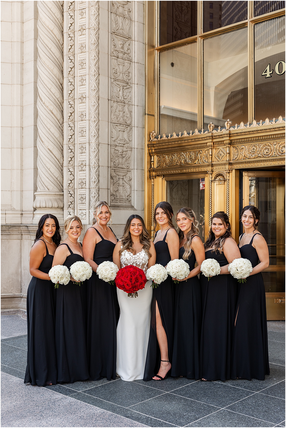 bride in corset style wedding dress holding red rose bouquet with bridesmaids in black dresses and holding white rose bouquets