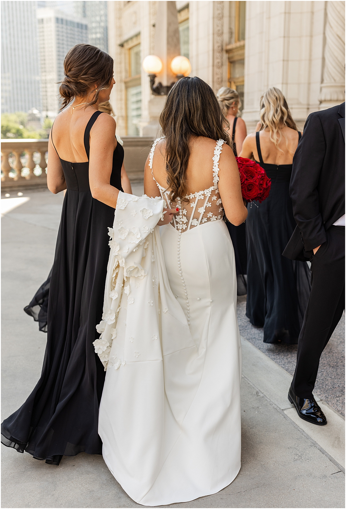 bridesmaid in black dress hold back of bride's dress as they walk
